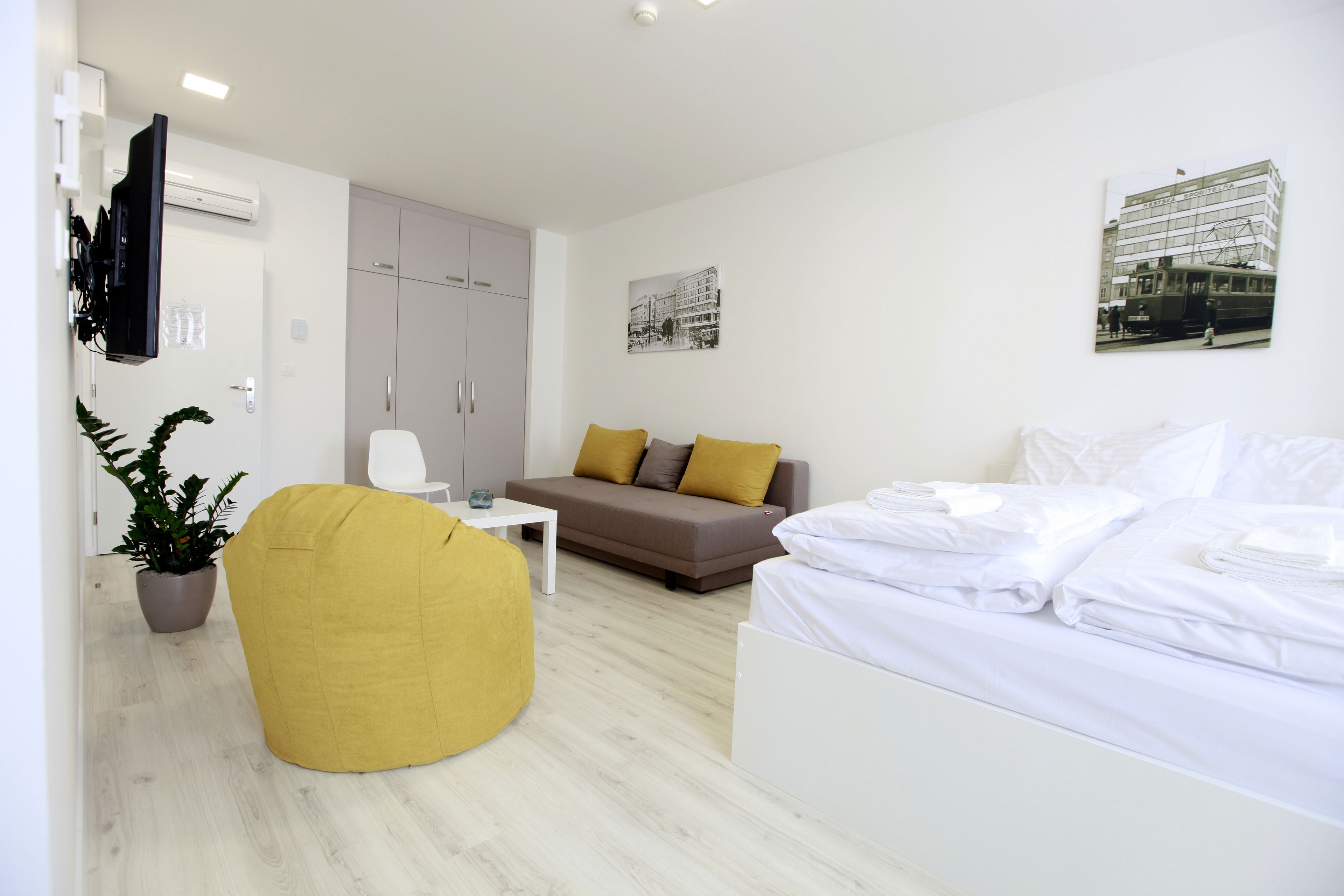 A modern hotel room with a bed, a sofa, a yellow bean bag, a small table, and a potted plant.