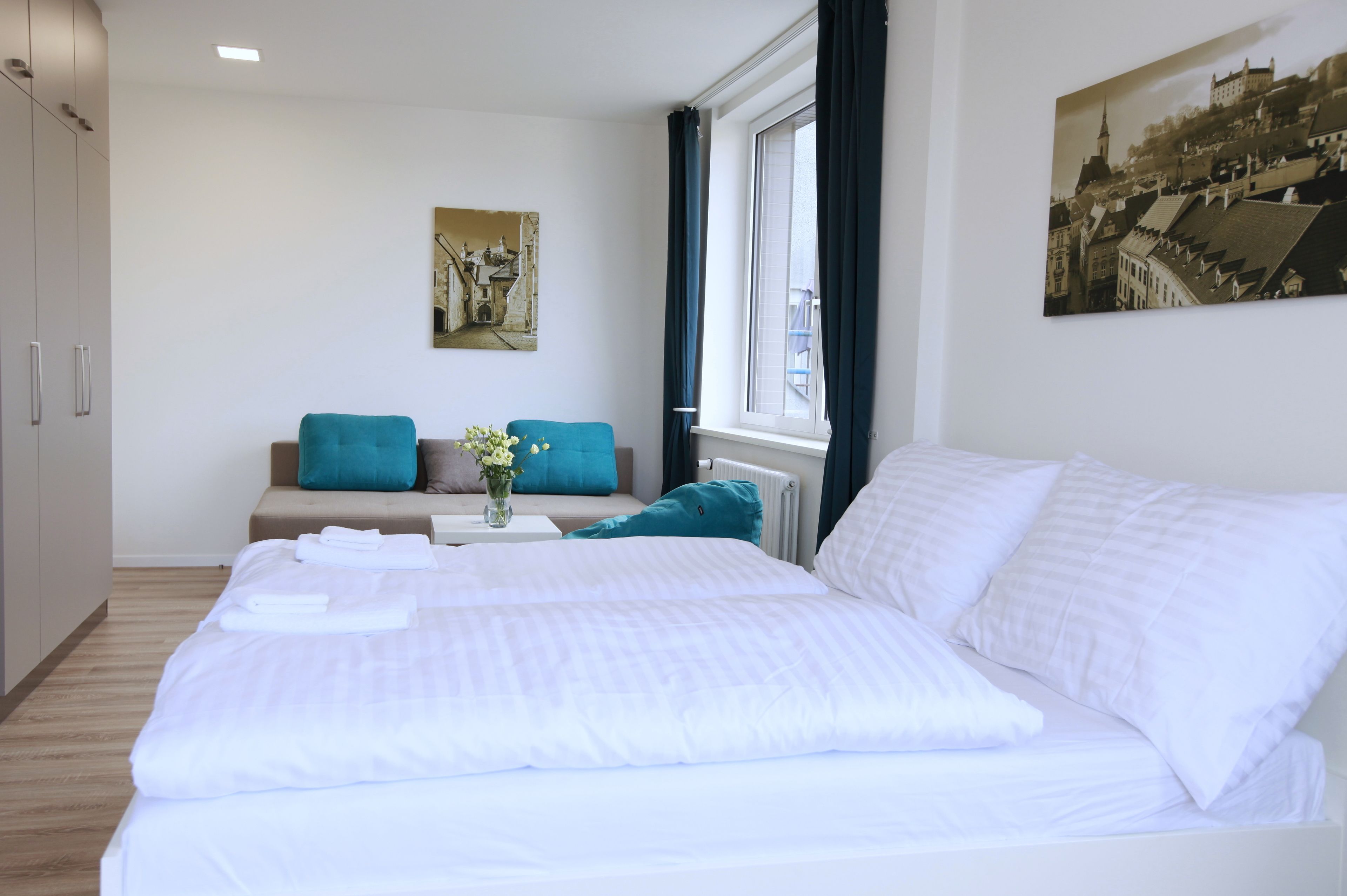 A hotel room with a large bed, a sofa with teal cushions, a small table with a vase of flowers, and a cityscape painting on the wall.