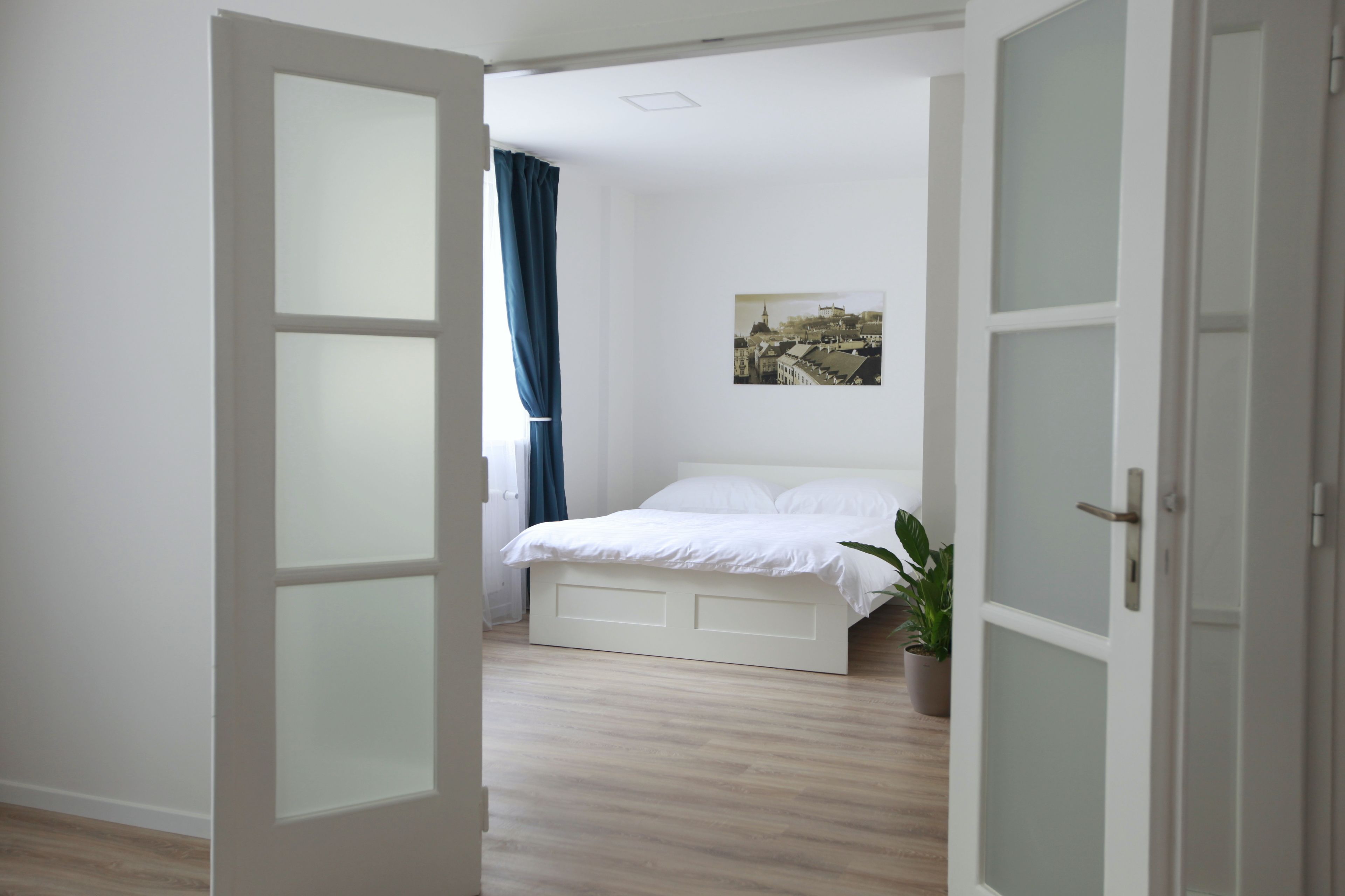 A bedroom with a bed, a potted plant, and a cityscape painting on the wall.