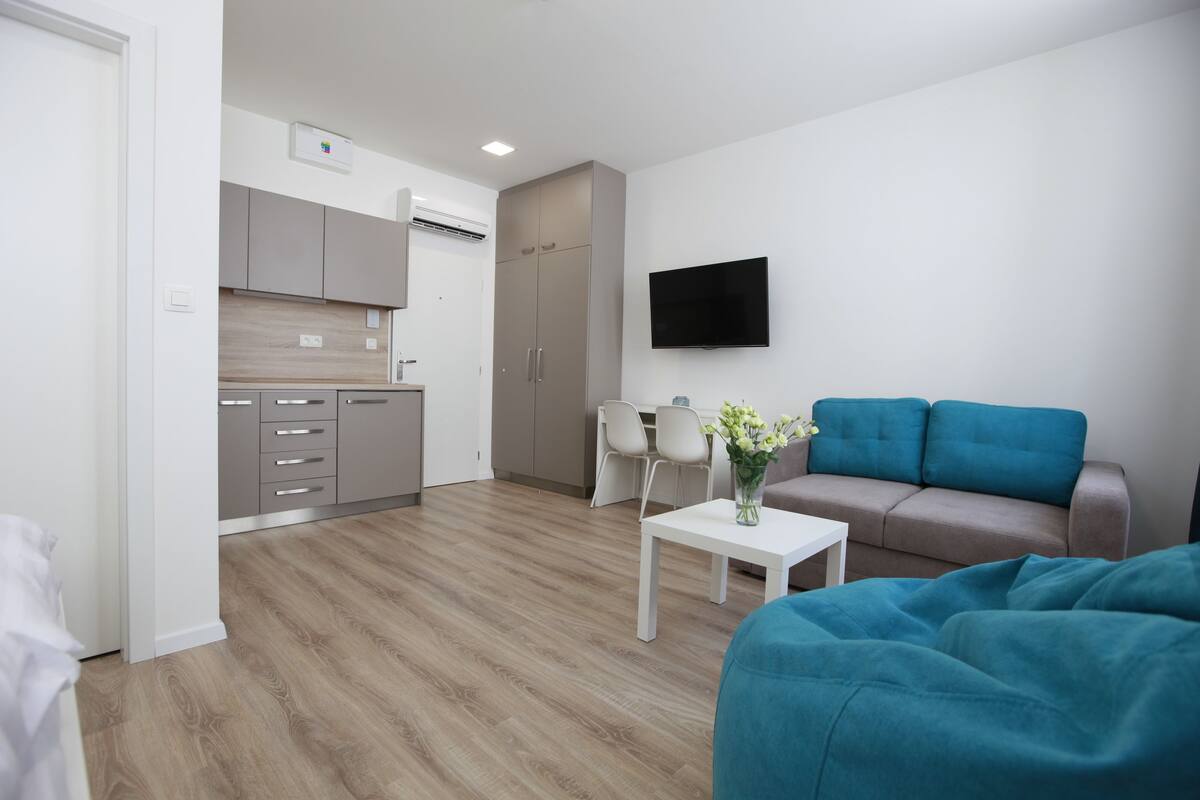 Ein modernes Wohnzimmer mit einer Couch, einem Couchtisch mit einer Vase, einem an der Wand montierten Fernseher und einer Küchenzeile im Hintergrund.