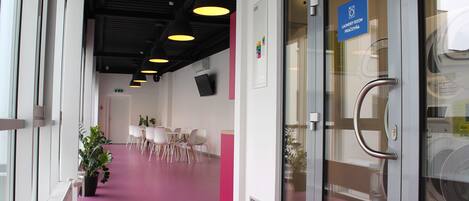 Un pasillo con alfombra rosa que conduce a una habitación con una puerta de vidrio que indica “Lavandería”. Se ven sillas y una mesa al fondo, y una planta en maceta a la izquierda.