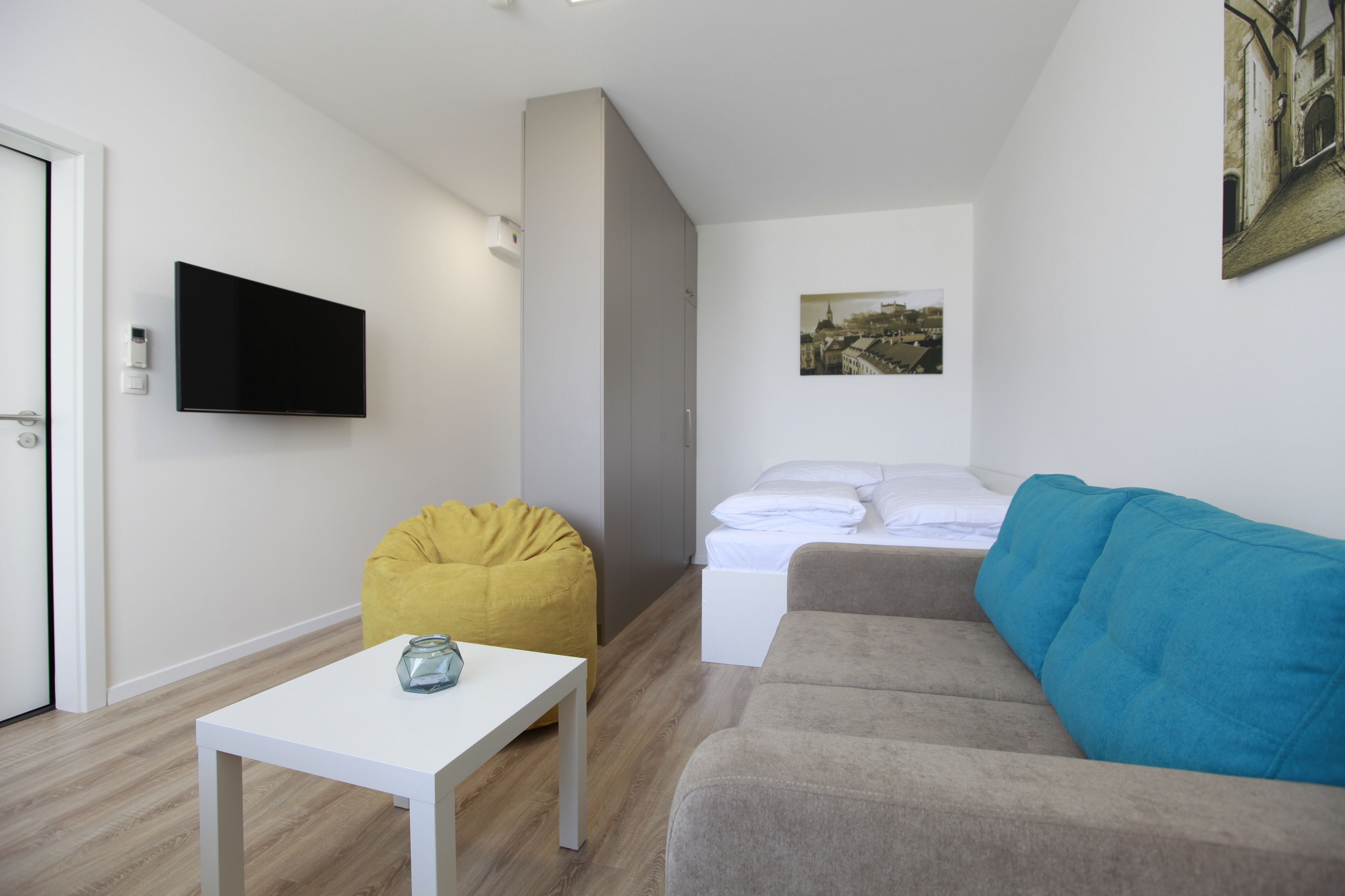 A modern, minimalistic room with a white table, a sofa, a bed, and a mounted TV.