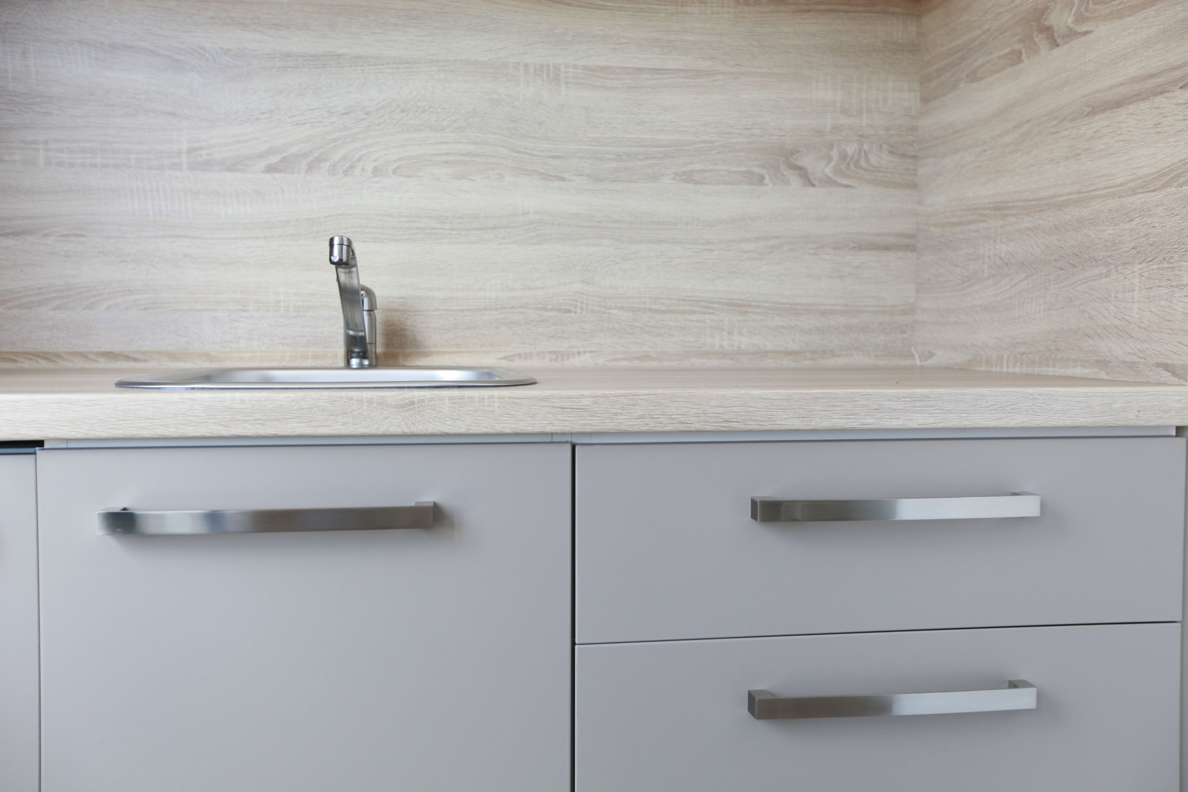A kitchen with a modern faucet, a light wood backsplash, and grey cabinetry with silver handles.