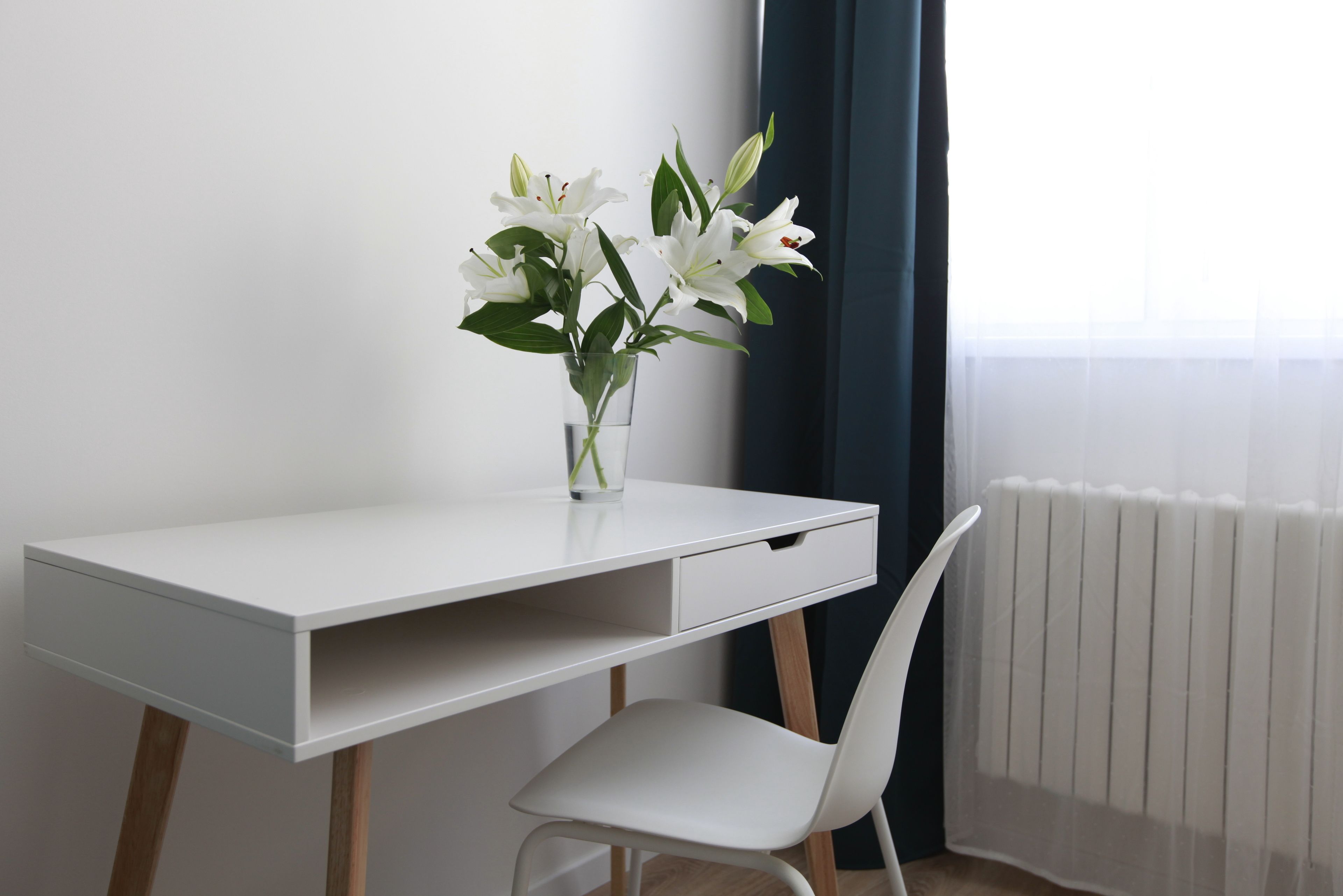 A white desk with a vase of white flowers, a white chair, and a window with a view of a building.