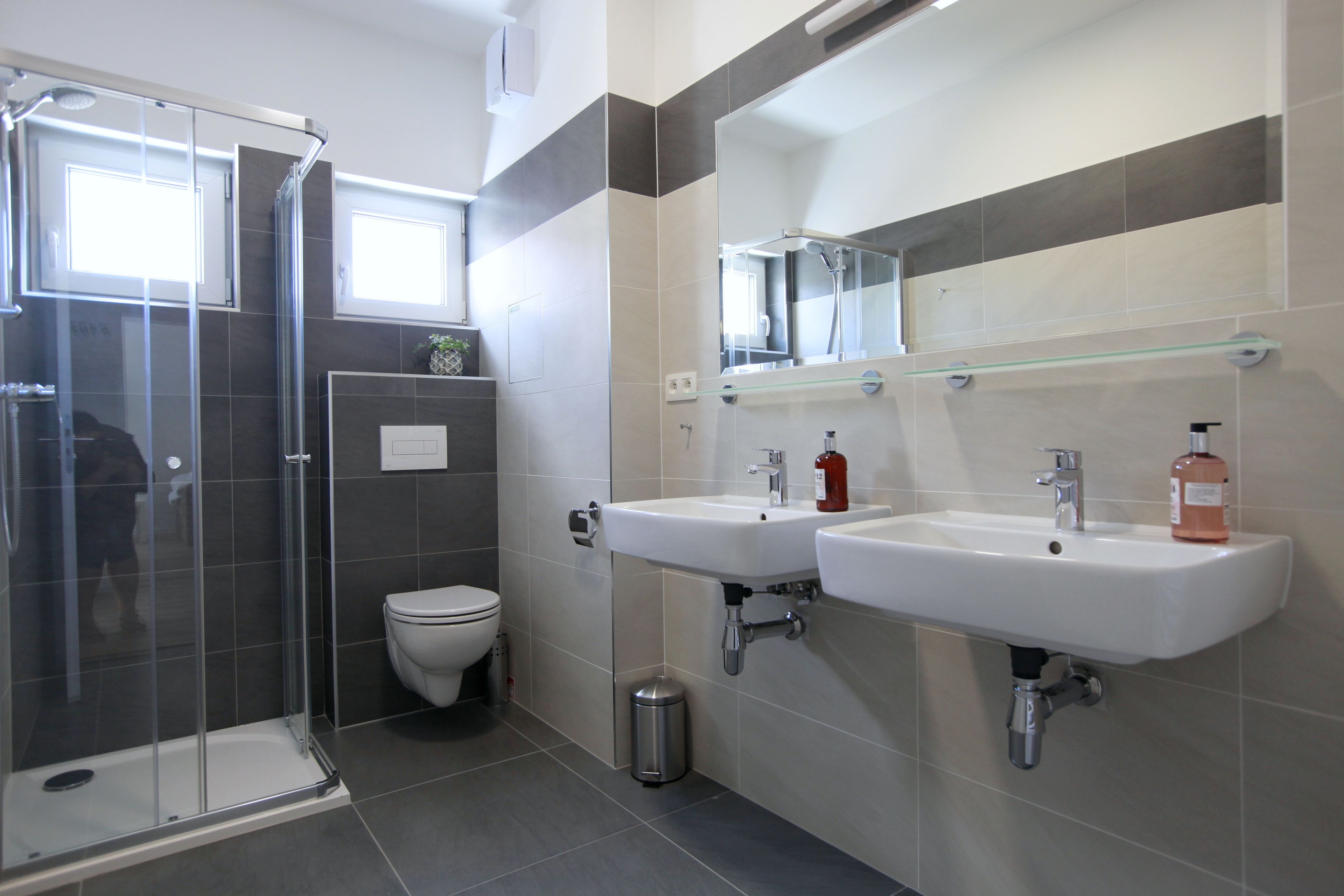Een moderne badkamer met een douchecabine van glas, een toilet en twee wastafels met een spiegel erboven.