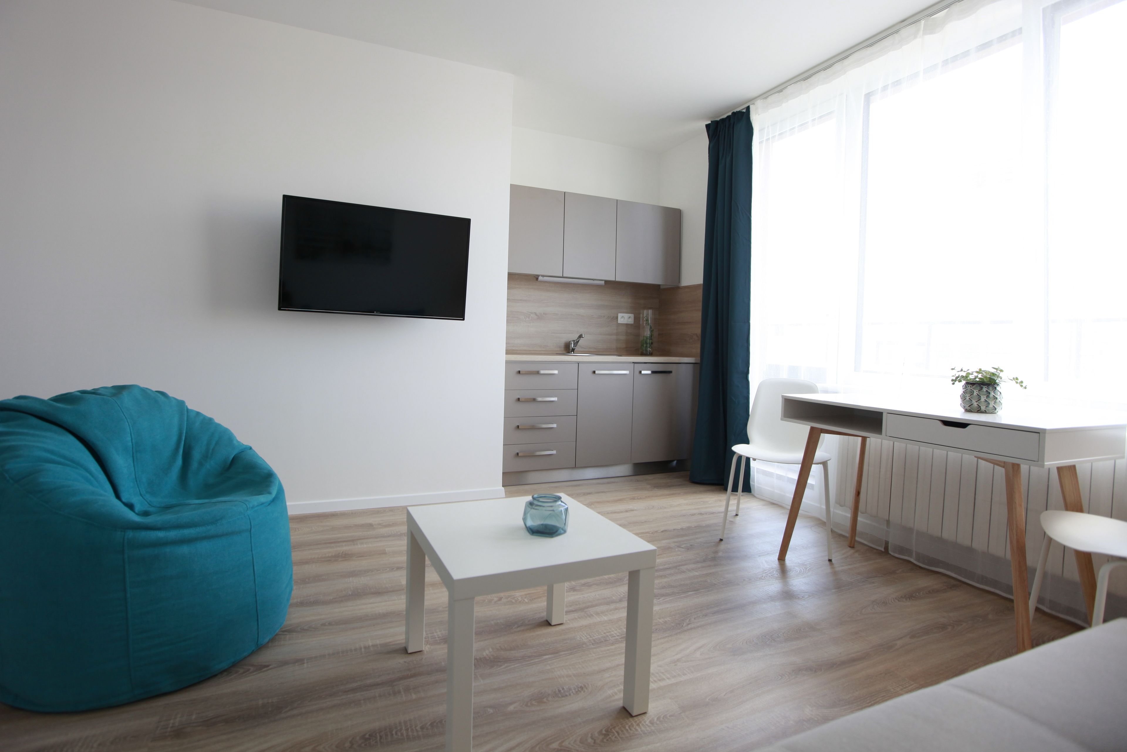 Una sala moderna con un sillón de peluche color turquesa, una mesa de centro blanca, un televisor de pantalla plana y una zona de cocina con ventana y cortinas.