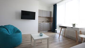 Una sala moderna con un sillón de peluche color turquesa, una mesa de centro blanca, un televisor de pantalla plana y una zona de cocina con ventana y cortinas.