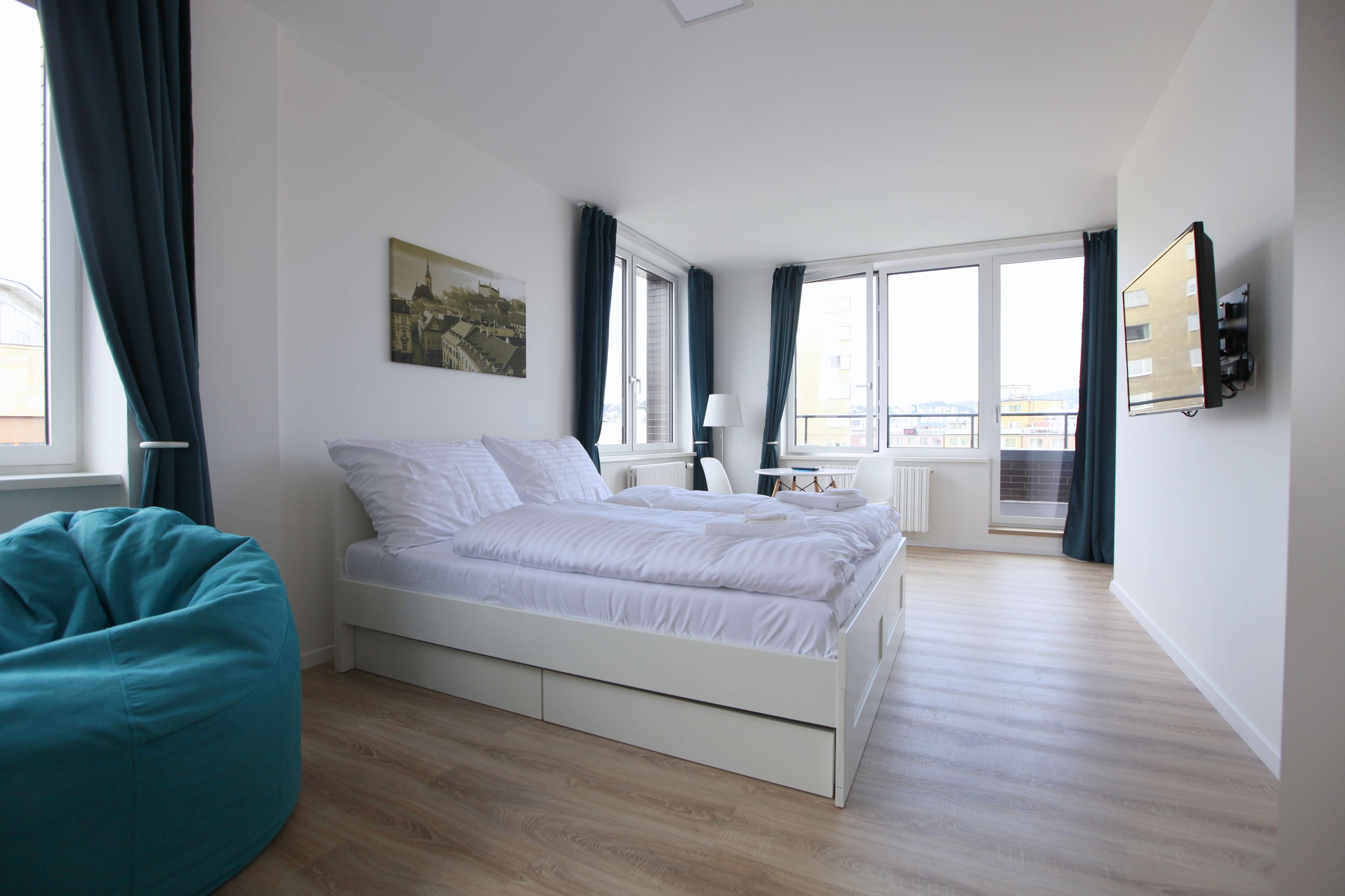 Un dormitorio moderno con cama, un sillón de peluche color turquesa, un escritorio y una ventana con cortinas.