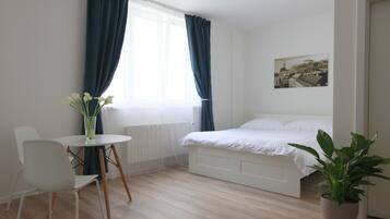Et soveværelse med seng, et lille bord med stole, en potteplante og et vindue med gardiner.