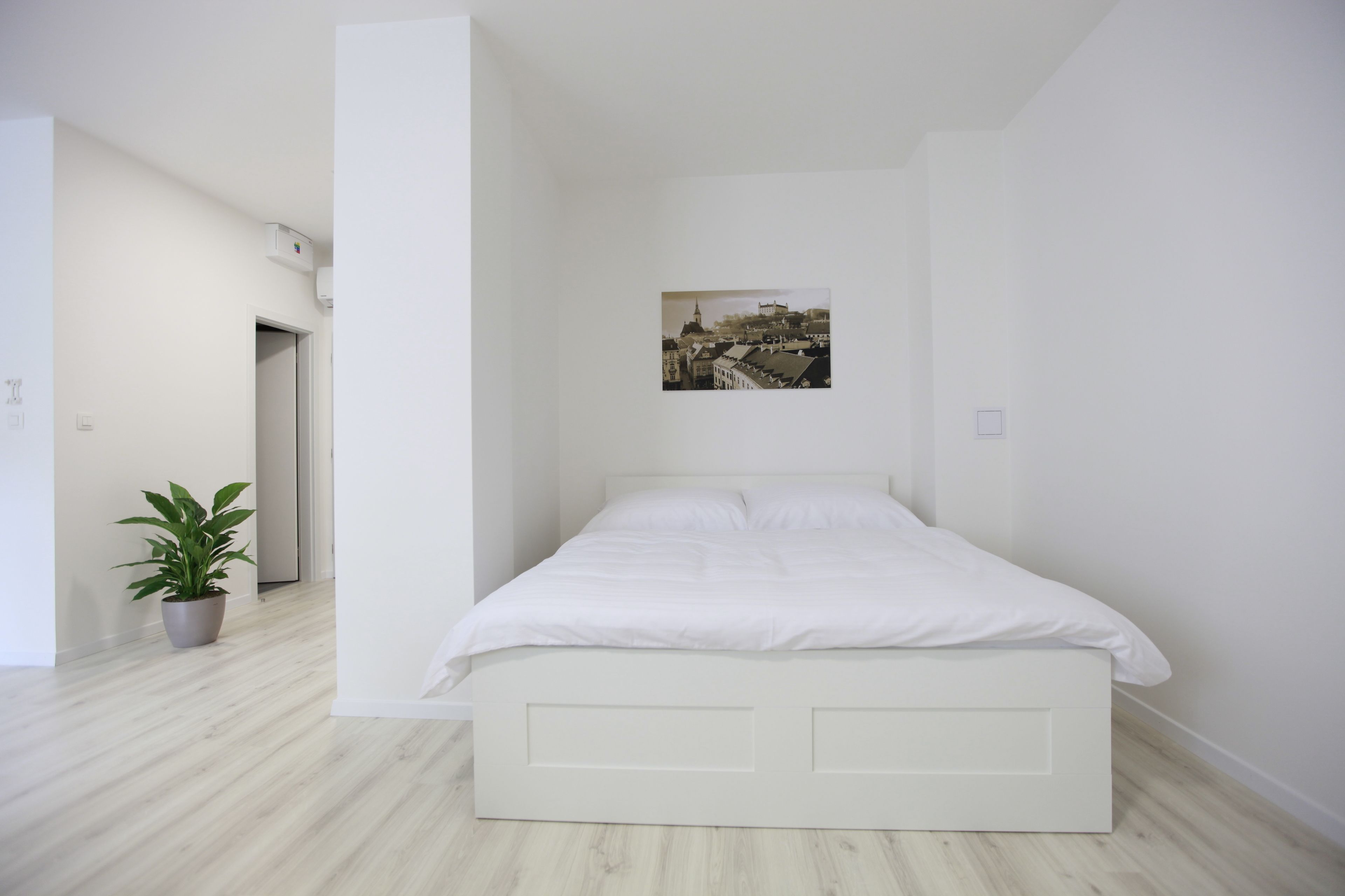A bedroom with a bed, a potted plant, and a cityscape photo on the wall.