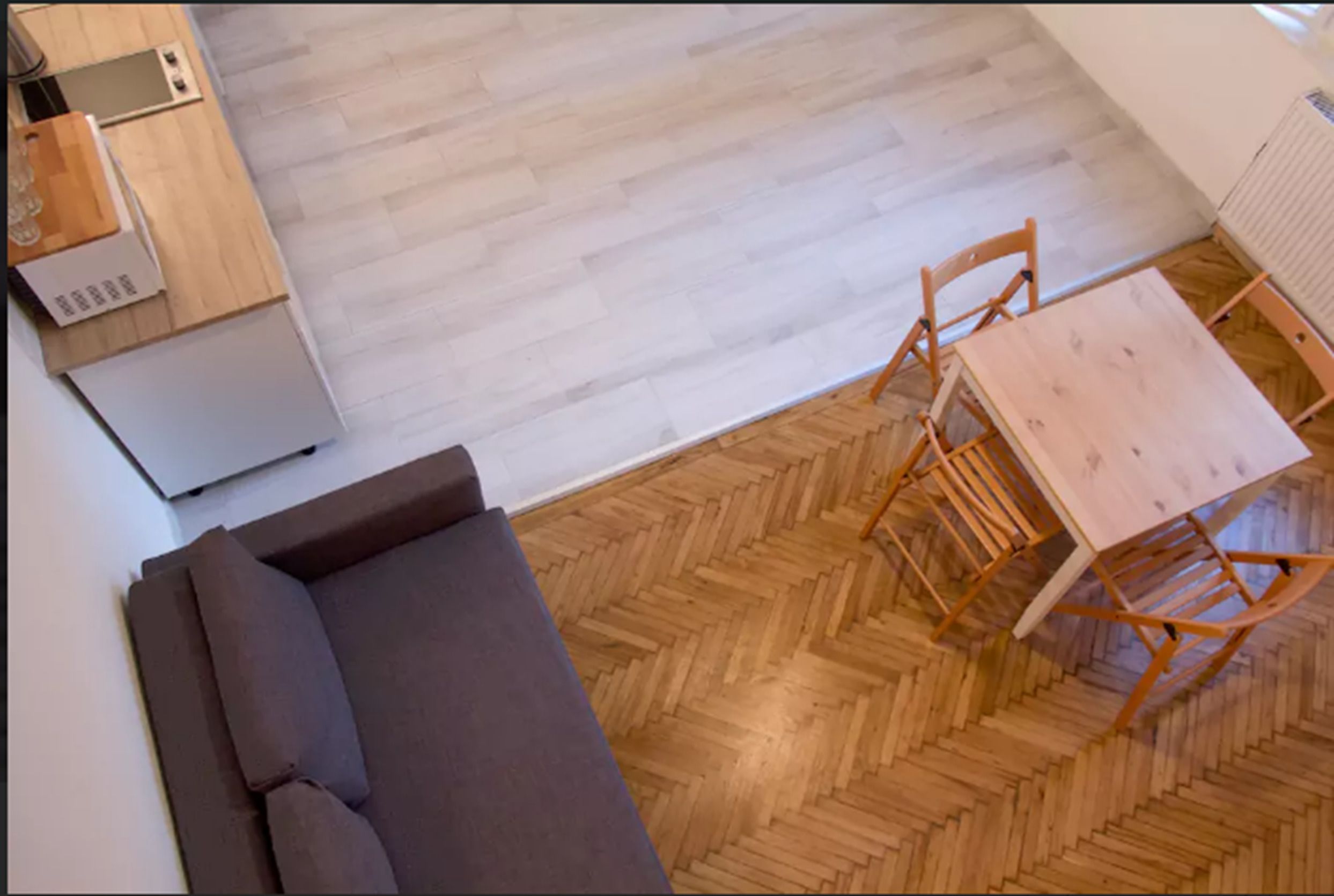 A small, modern room with a wooden herringbone floor, a gray sofa, a wooden dining table with chairs, and a kitchen area with a refrigerator and microwave.