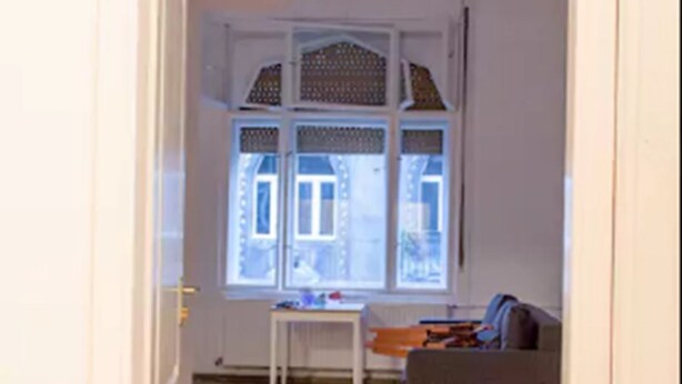 Une pièce avec un parquet en chevrons, une porte blanche, une fenêtre ornée de rideaux à motifs et un canapé gris.
