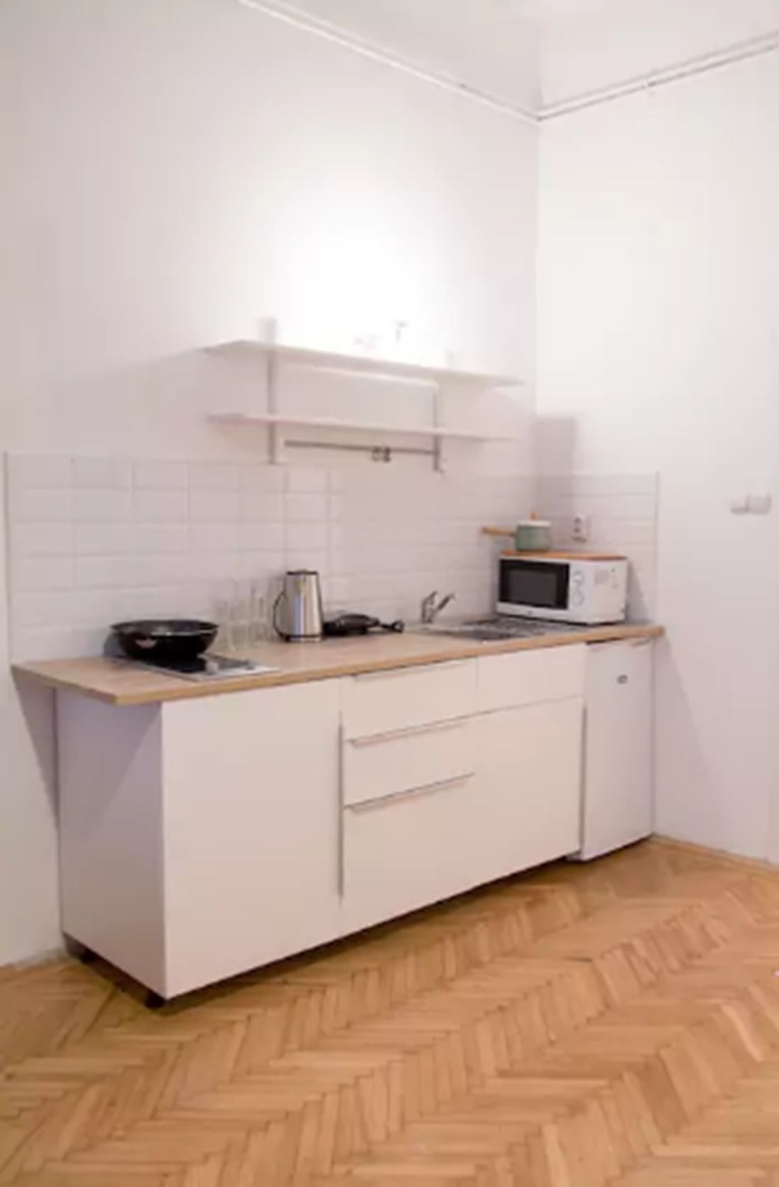 A small kitchenette with white cabinets, a microwave, a kettle, and a wooden countertop.