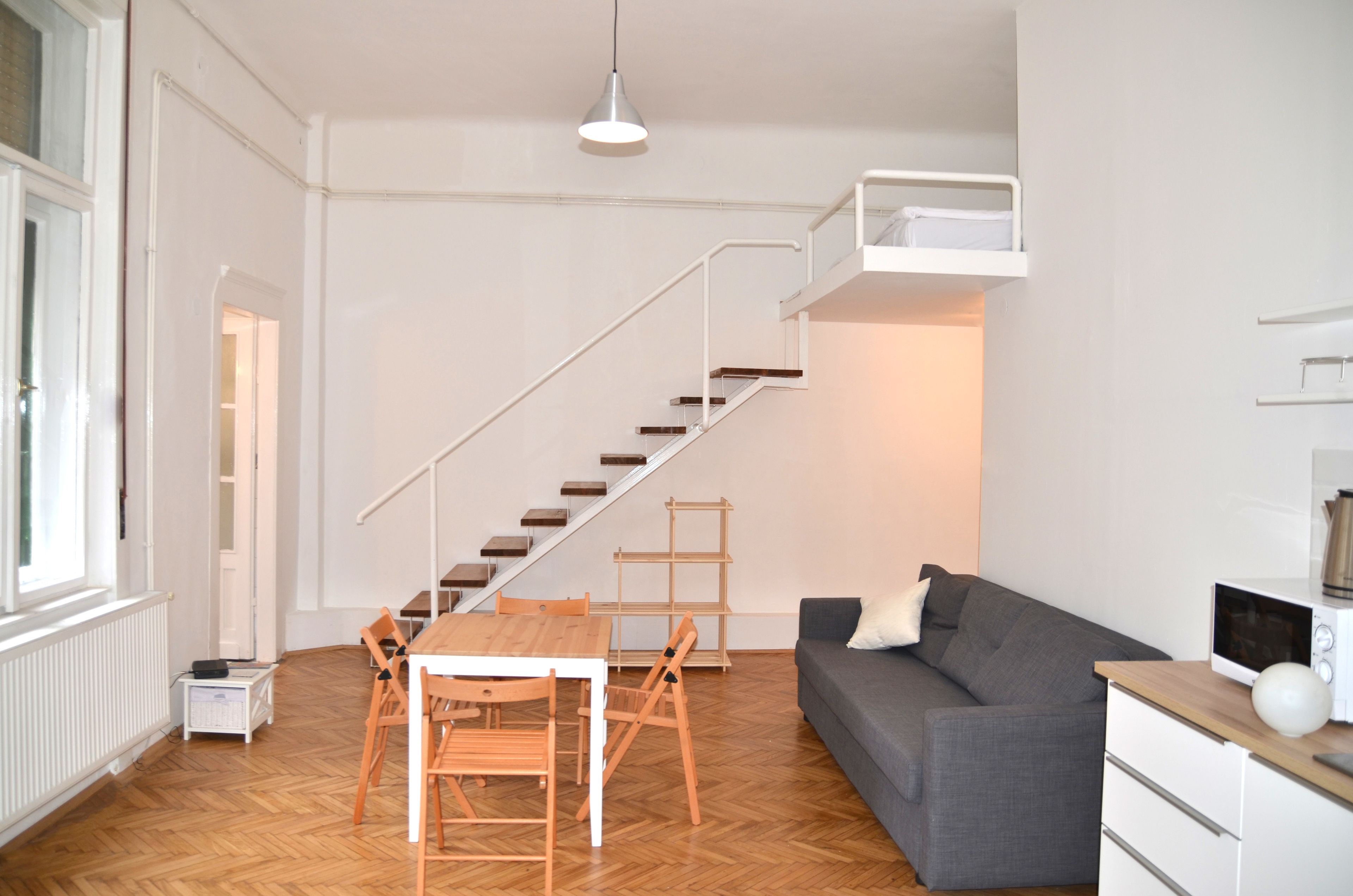 A modern living room with a wooden floor, white walls, a grey sofa, a wooden dining table with chairs, a white staircase, and a white radiator.