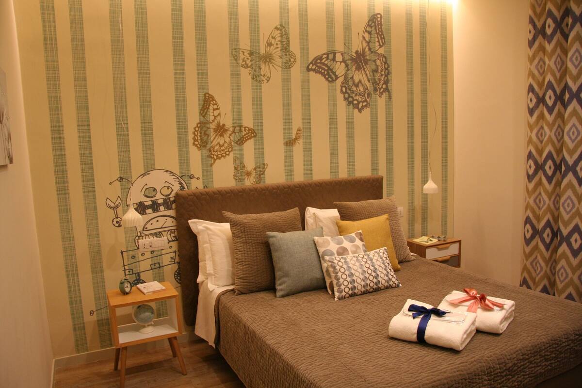 A neatly made bed with a brown quilt, multiple pillows, and two neatly folded towels with ribbons. There is a small bedside table with a lamp and a decorative item. The wall features a striped pattern with butterfly designs.