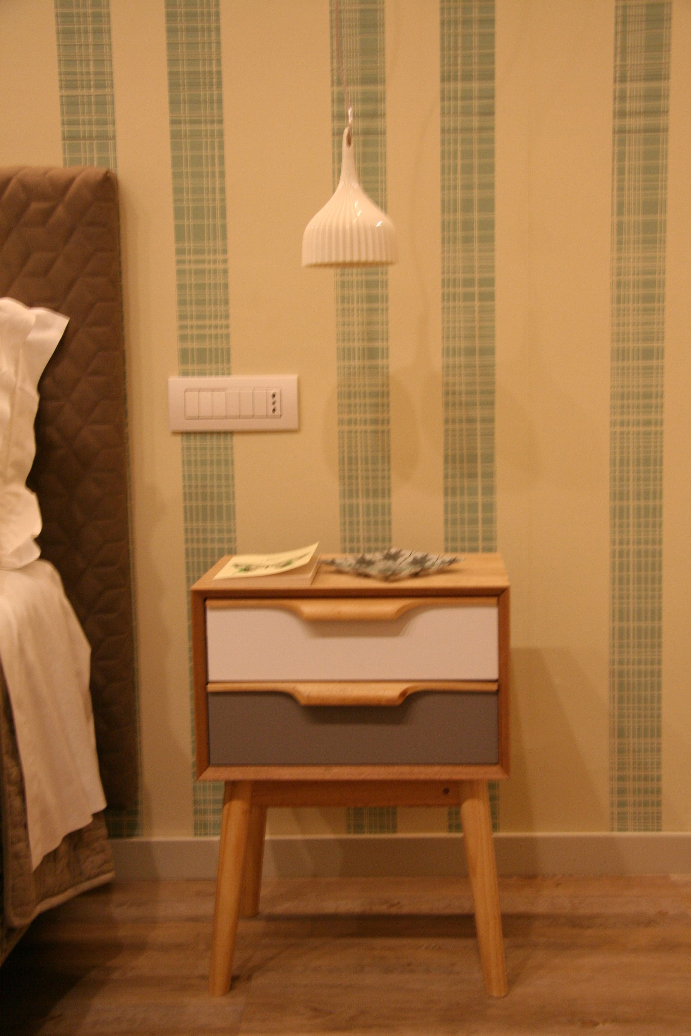 Een klein houten nachtkastje met een lade, een witte lamp en een bruine gewatteerde stoel.