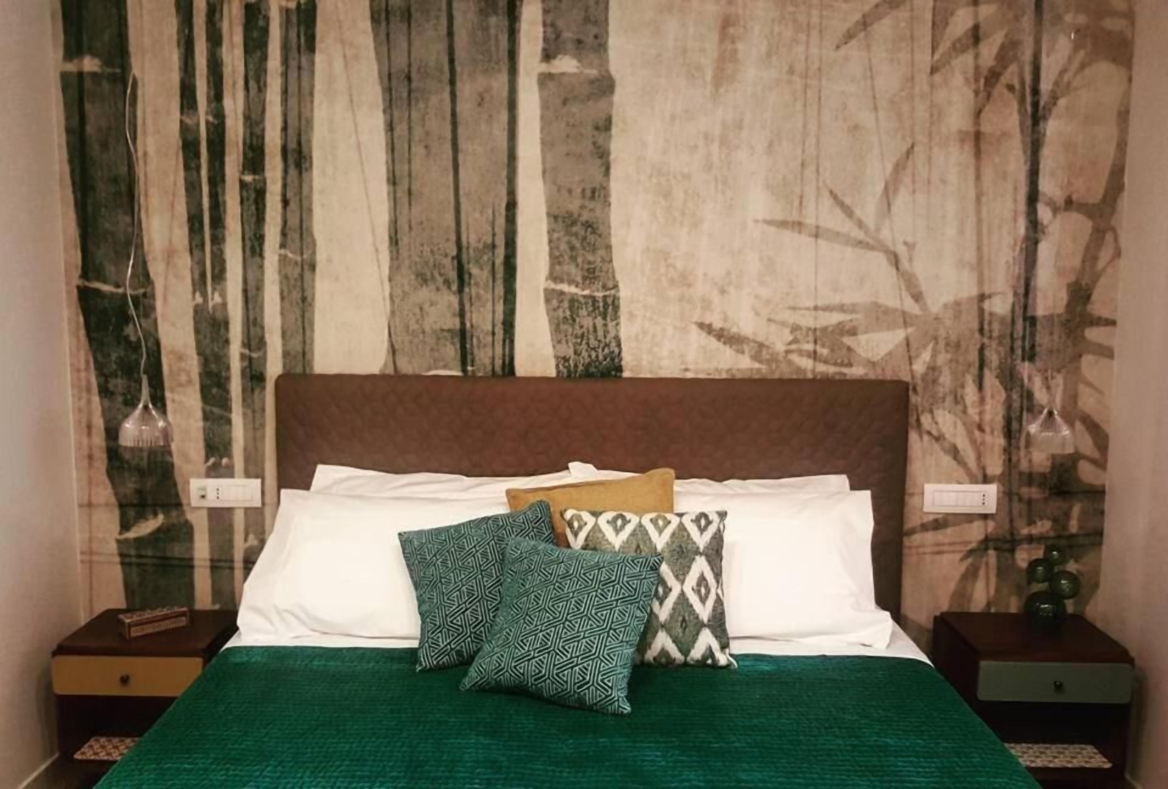 A bed with a green bedspread, white pillows, and patterned pillows. There are two nightstands with lamps and a wall with a bamboo pattern.