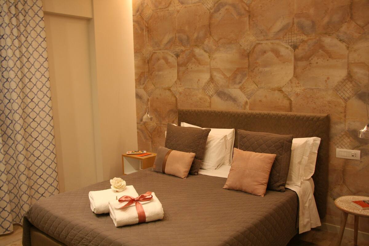 A neatly made bed with a brown quilt, several pillows, and two neatly folded towels with a ribbon.