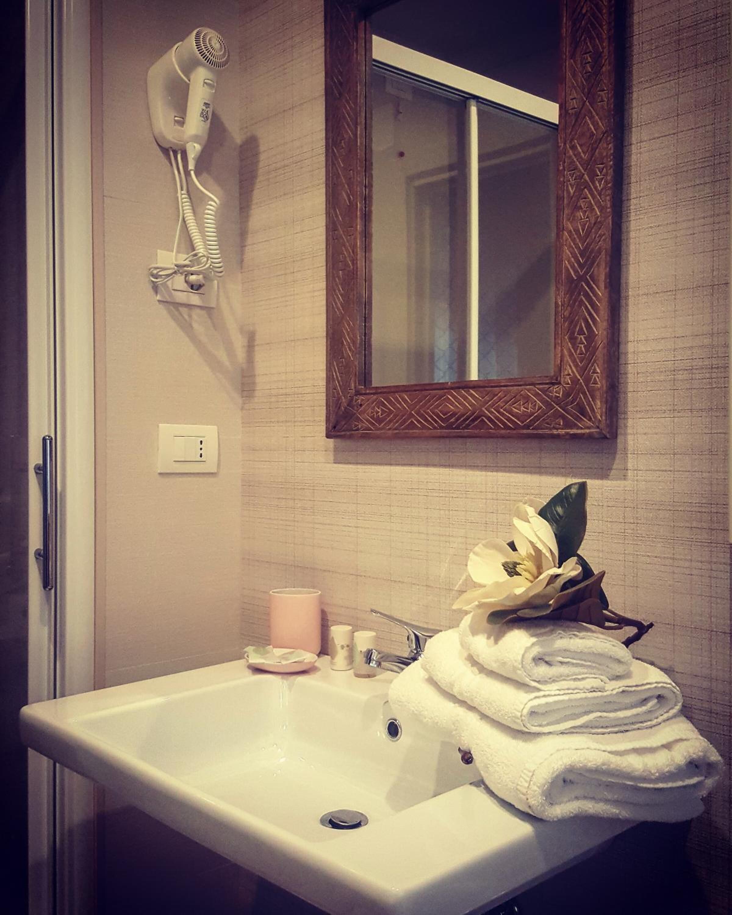 Een badkamer met een witte wasbak, een aan de muur bevestigde haardroger, een spiegel met decoratieve lijst en een handdoekrek met opgevouwen handdoeken.