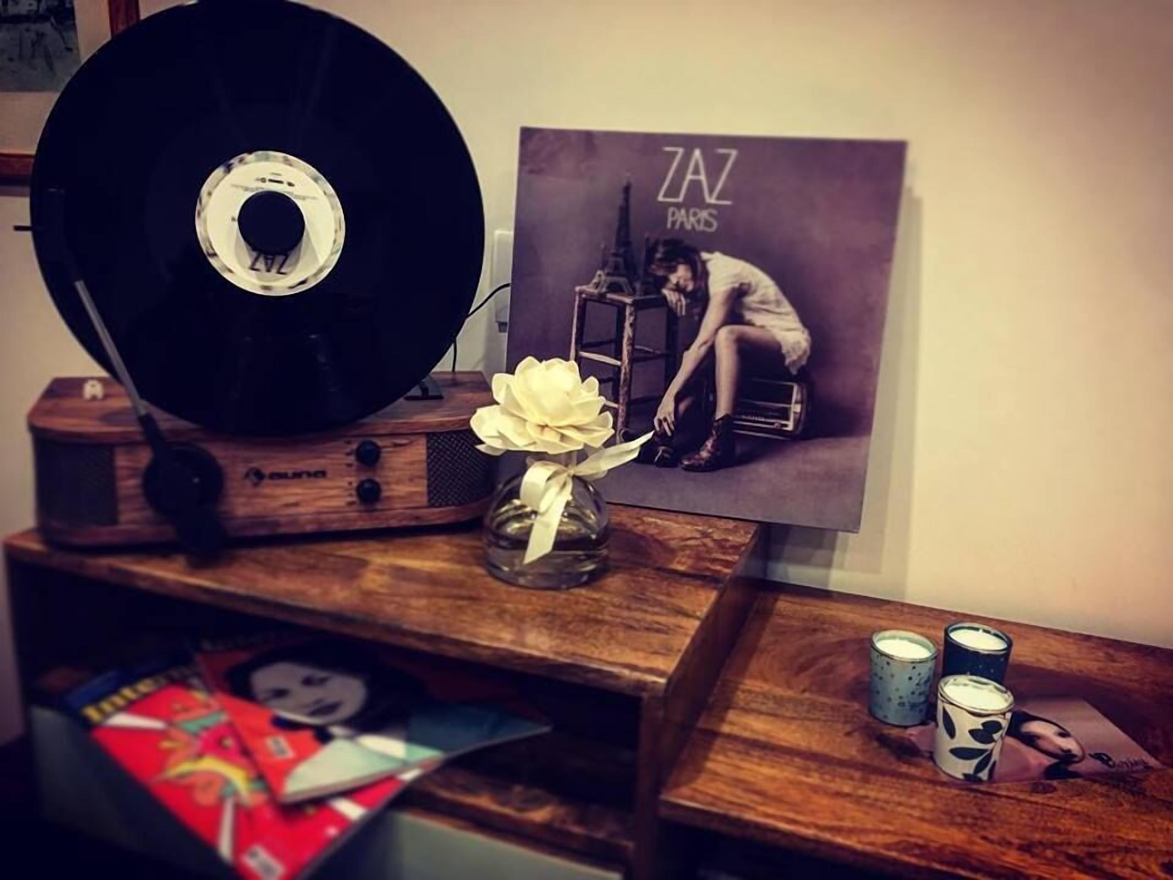 A wooden table with a vinyl record, a vase with a white flower, a poster of a woman sitting on a ladder, and a few cans.