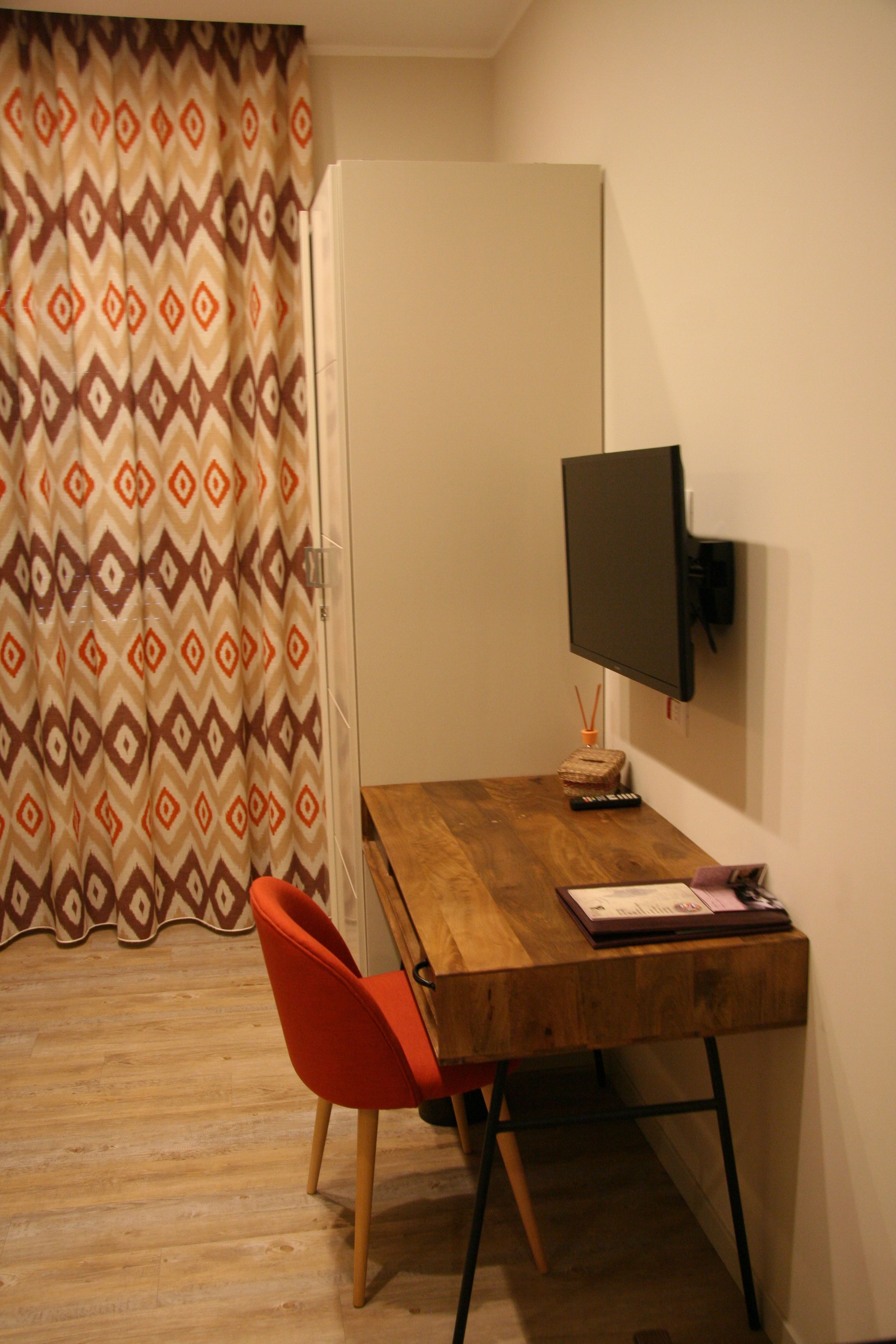 A small room with a wooden desk, a single orange chair, a television mounted on the wall, and a patterned curtain.