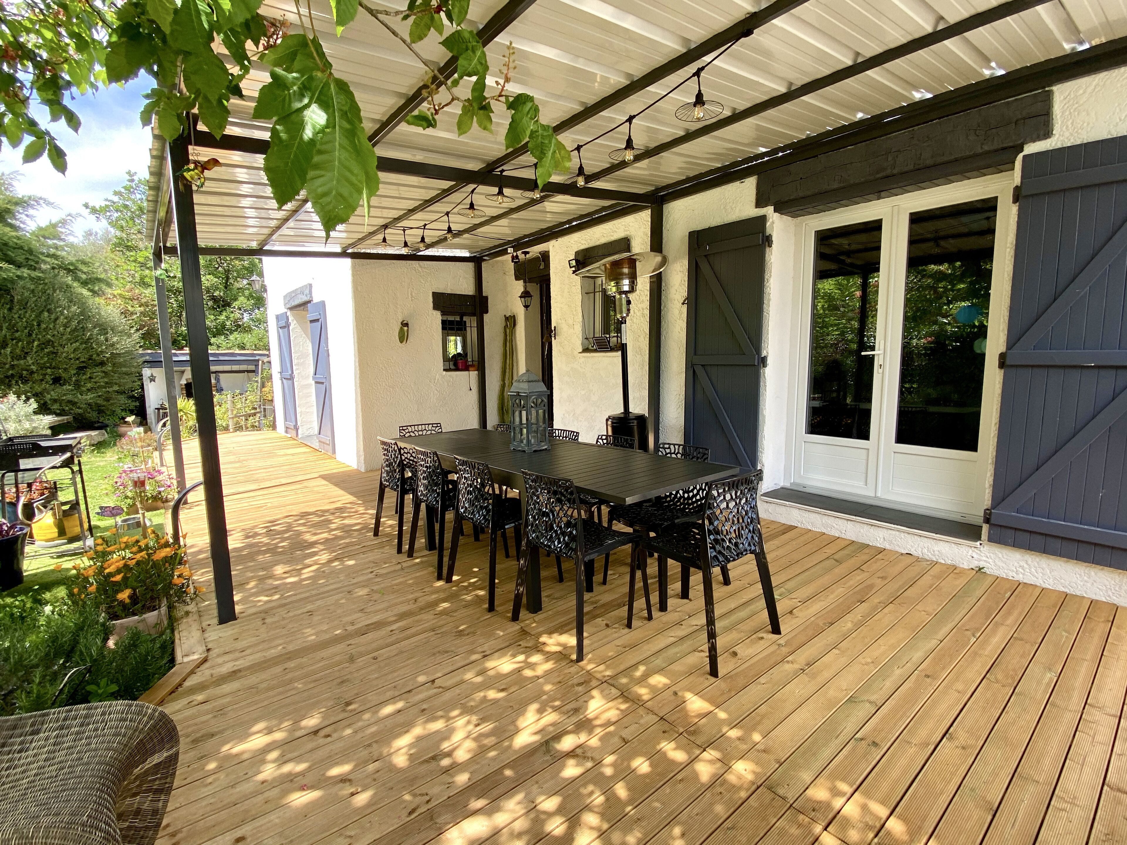 An outdoor patio with a wooden deck, a table with chairs, and a pergola with hanging lights.