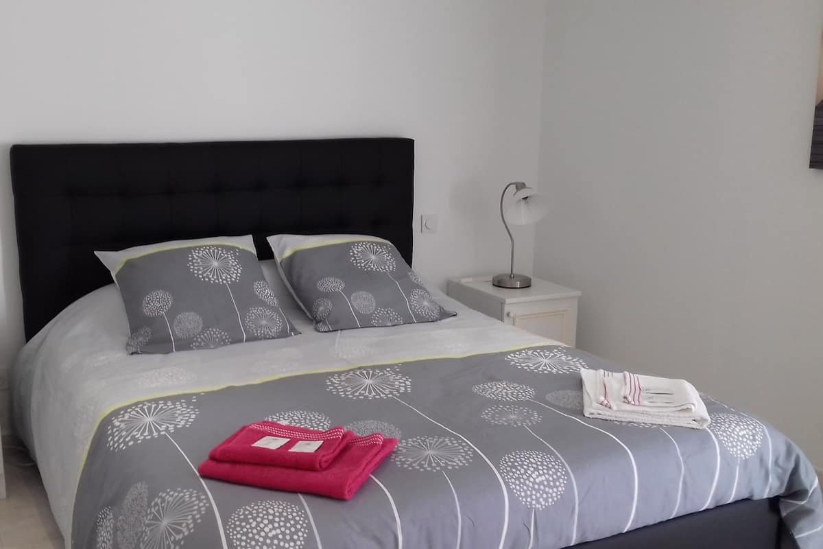 A neatly made bed with a grey and white floral bedspread, two matching pillows, and a red folded towel on the bed. There are two bedside tables with lamps on either side of the bed.