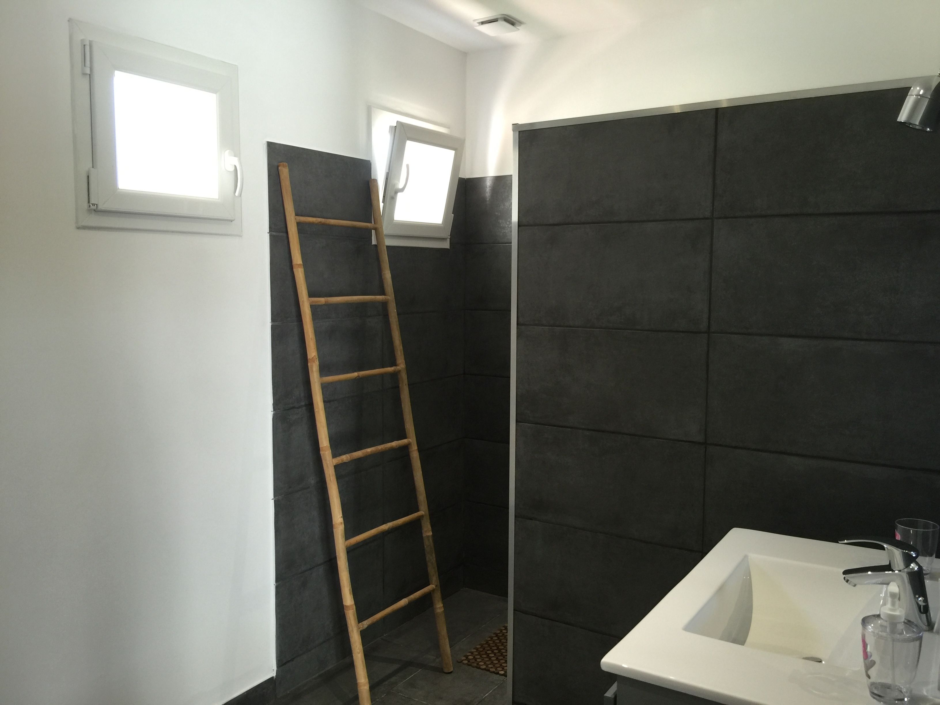 A bathroom with a white sink, dark tiled walls, a wooden ladder, and a window.