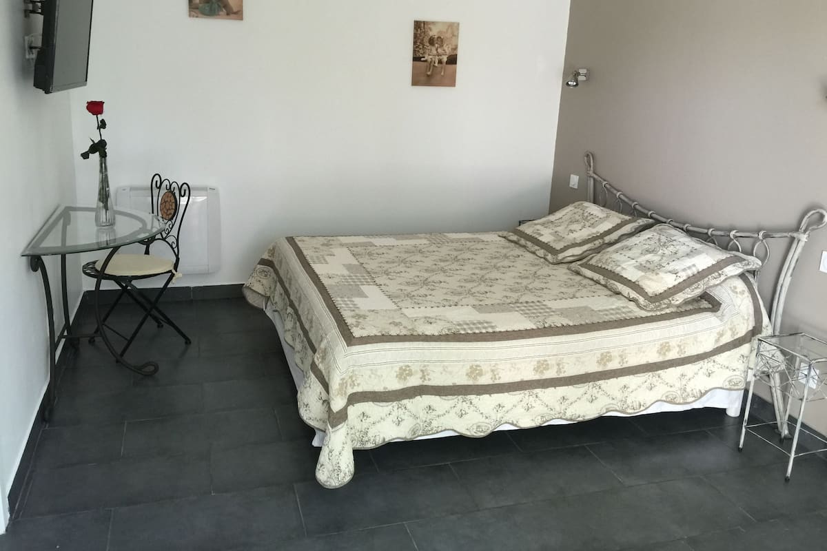 A bedroom with a bed, a small table with a chair, a vase with a rose, and two wall-mounted pictures.