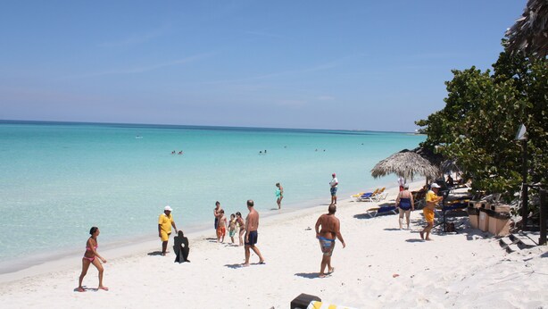 Eine Strandansicht mit klarem türkisfarbenem Wasser, Menschen, die am Sand entlanggehen, und Strandsonnenschirmen.