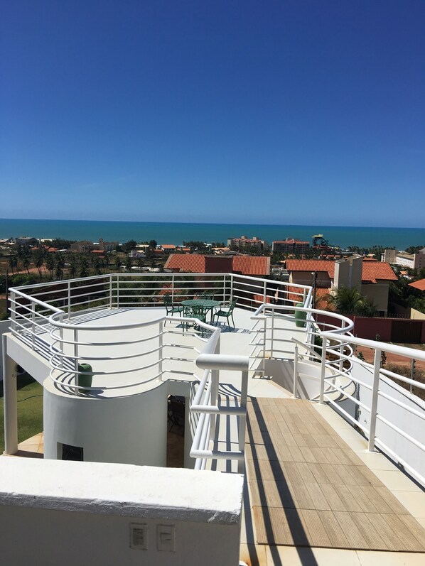 A rooftop terrace with a white railing, a wooden deck, and a view of the sea and buildings in the distance.