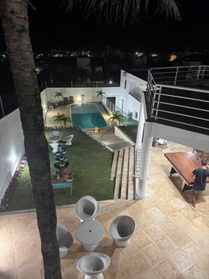 A modern outdoor pool area with a staircase leading to a covered deck, surrounded by palm trees and a well-maintained lawn.