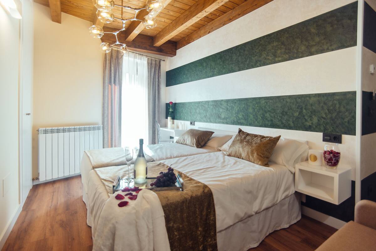 Habitación de hotel con dos camas, techo de madera, radiador, mesita de noche con un florero y otra mesita con una lámpara.