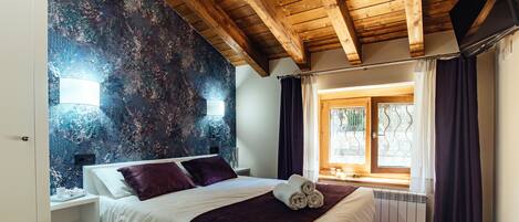 A bedroom with a wooden ceiling, a large bed with white bedding, a purple throw blanket, and a window with curtains.
