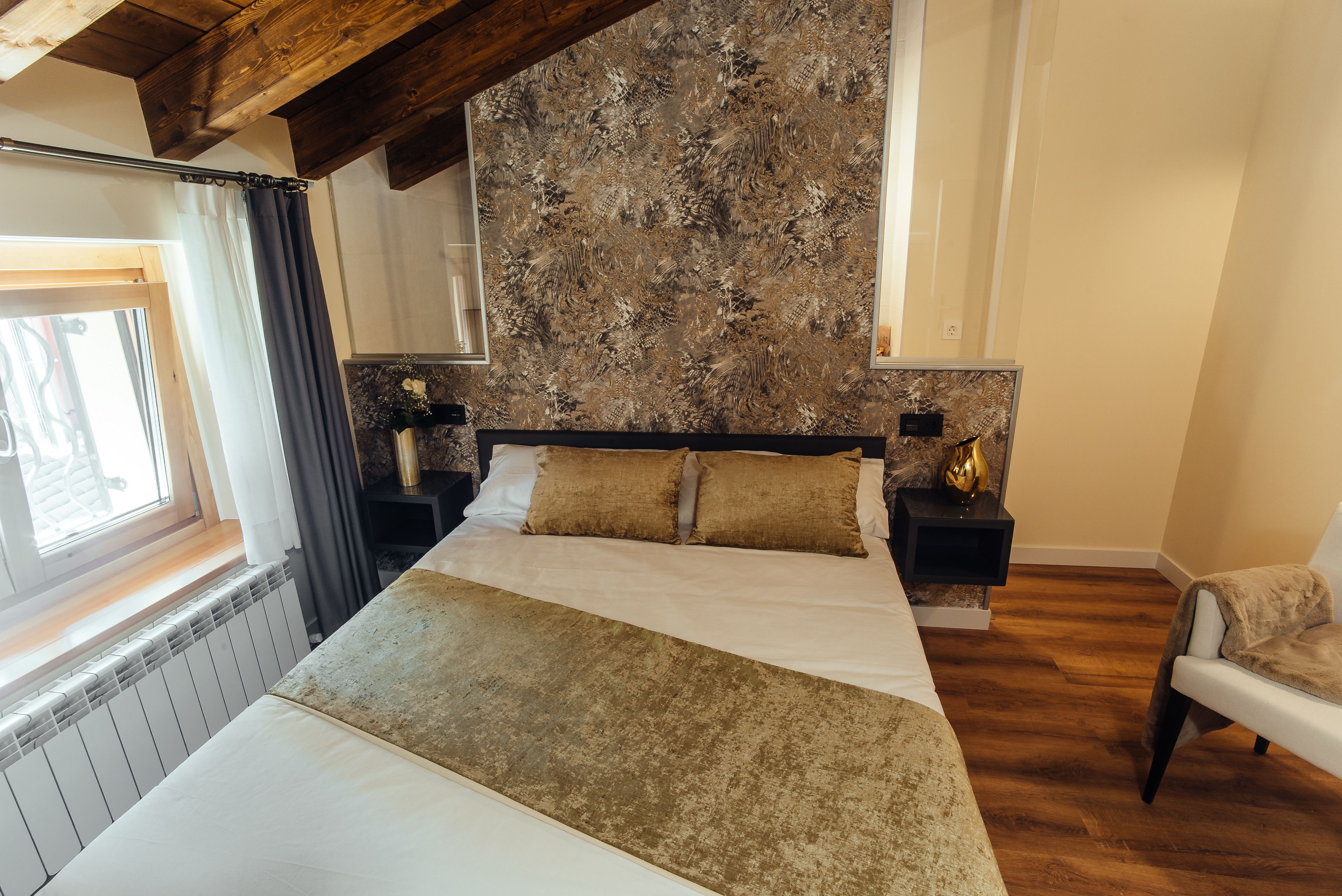 A bedroom with a large bed, wooden beams, a stone accent wall, a window with curtains, and a nightstand with a lamp.