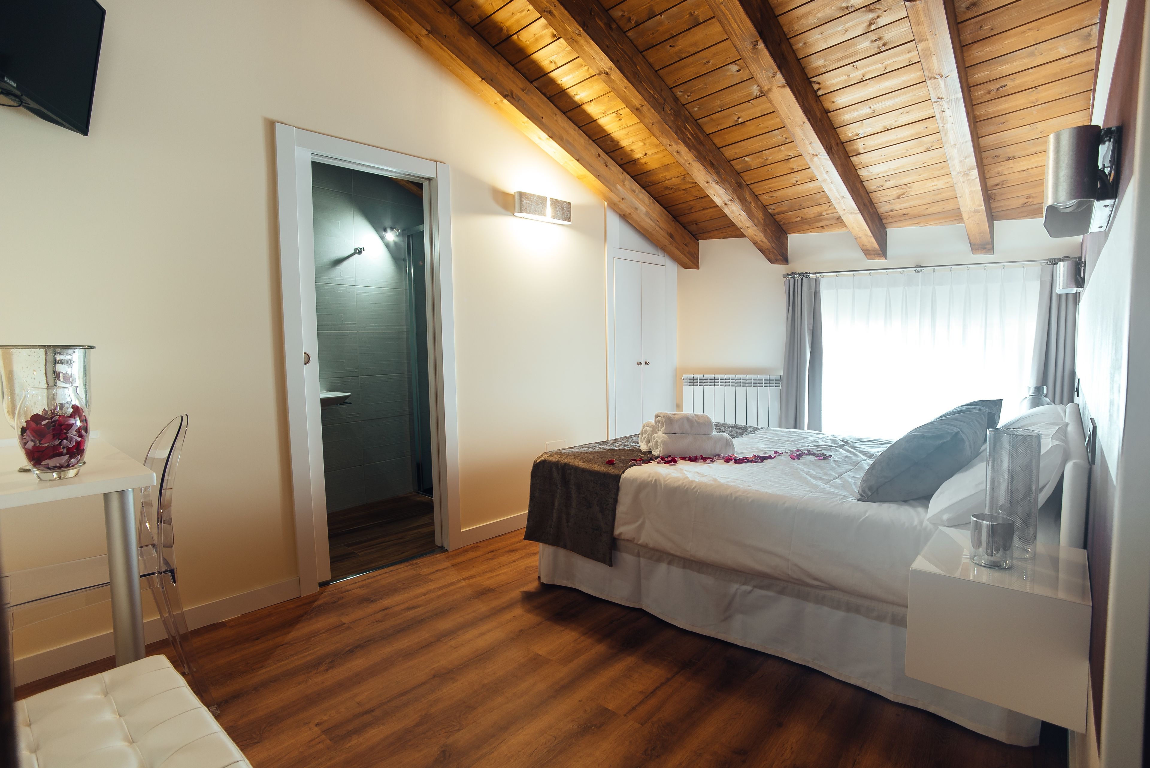 Camera da letto con soffitto in legno, un letto con lenzuola bianche, un comodino con un bicchiere e una porta che dà su un'altra stanza.