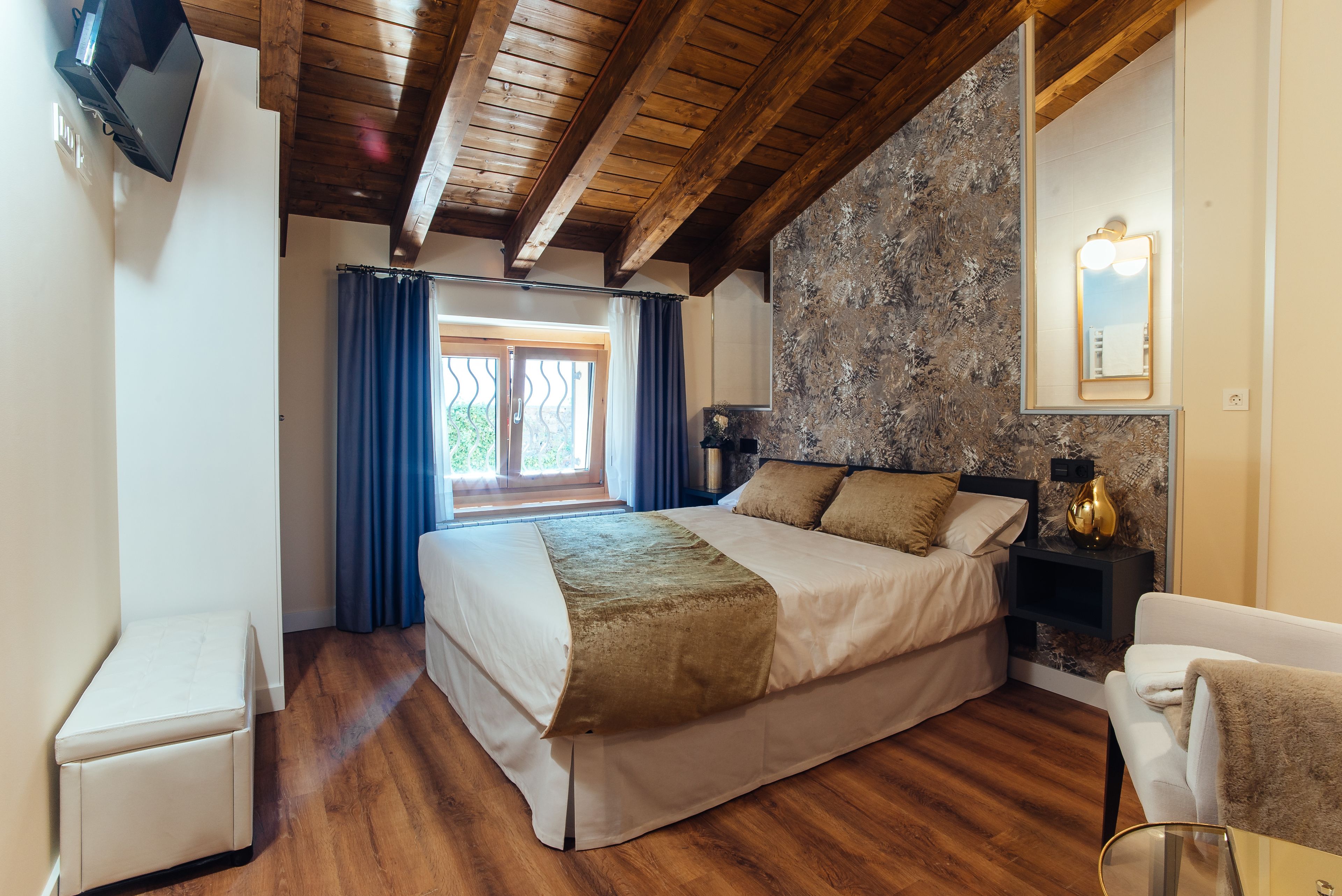 Un dormitorio con una cama grande, vigas de madera en el techo, una pared de piedra, una mesita de noche y un televisor montado en la pared.