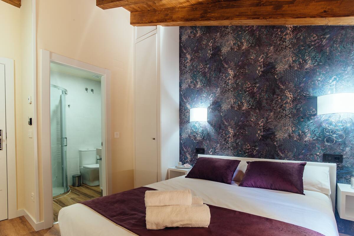 Habitación de hotel con cama, mesita de noche, pared con un mural texturizado, zona de ducha y puerta que da a otra habitación.