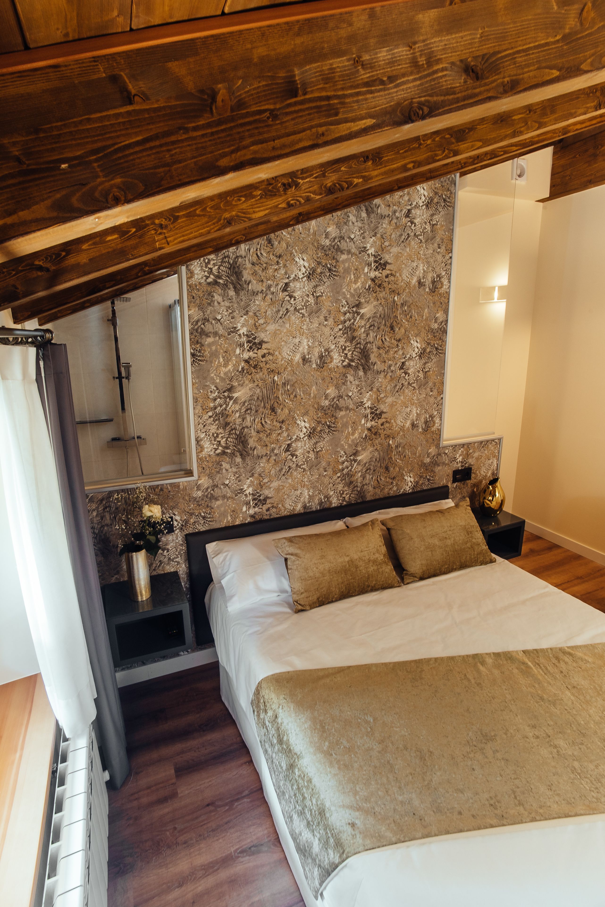 Un dormitorio con una cama grande, vigas de madera en el techo, pared con textura, una mesita de noche con un florero y un espejo.