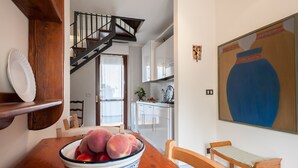 Un coin repas avec une table en bois, un saladier de fruits, un escalier, une cuisine et un grand tableau accroché au mur.