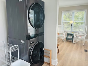 A laundry room with stacked washers and dryers, a white metal shelf, and a view of a porch with a rocking chair and a lamp. - Buckeye's At The Beach 3 Bedroom Home by RedAwning (Supply)