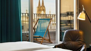 A hotel room with a bed, a leather chair, a blue hammock, and a view of a cathedral. - Motel One Köln-Neumarkt (Cologne)