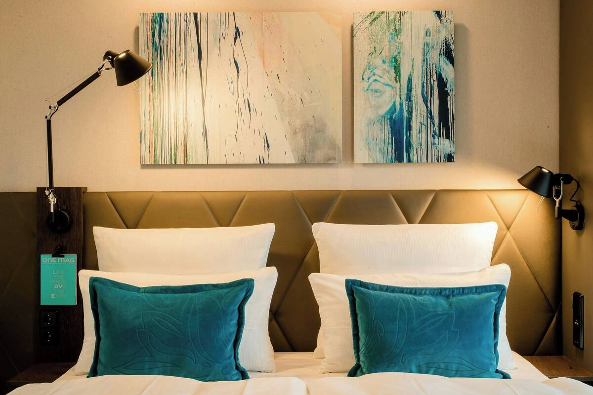 A hotel room with a tufted headboard, white bedding, teal pillows, and abstract artwork on the wall.