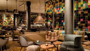 A modern lounge area with leather sofas, round wooden tables, and a colorful mosaic wall. - Motel One Köln-Neumarkt (Cologne)