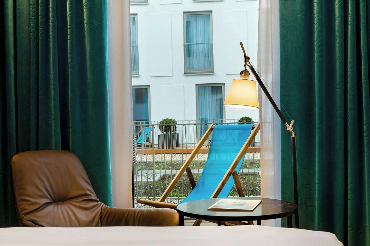 A hotel room with a bed, a brown leather chair, a small table with a book, a floor lamp, and a view of a building with windows and a balcony.