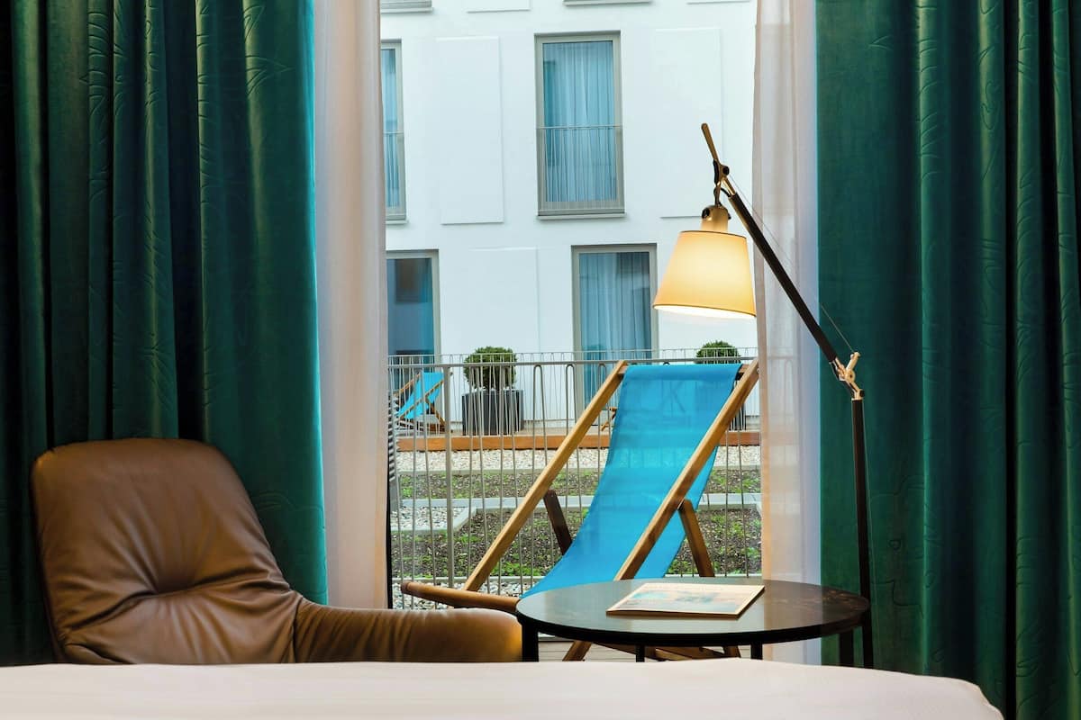 Habitación de hotel con una cama, un sillón de cuero marrón, una mesita con un libro, una lámpara de pie y vistas a un edificio con ventanas y un balcón.