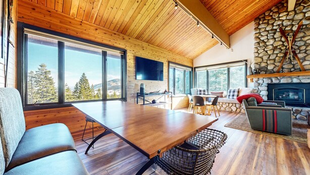 A spacious living area with a large wooden table, comfortable seating, a stone fireplace, and large windows offering a view of trees and mountains. - Bridges Townhome 5 Large, Remodeled Mountain View Townhome with Private Jacuzzi, Steps to Eagle Lodge by RedAwning (Mammoth Lakes)