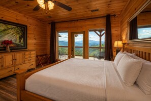 Une chambre dans une cabane en bois, avec un grand lit, un ventilateur de plafond, un tableau, une commode et un balcon offrant une vue sur les montagnes.