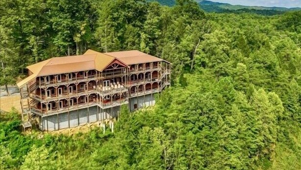 A large, multi-story wooden building with a balcony, surrounded by dense forest and mountains in the background.