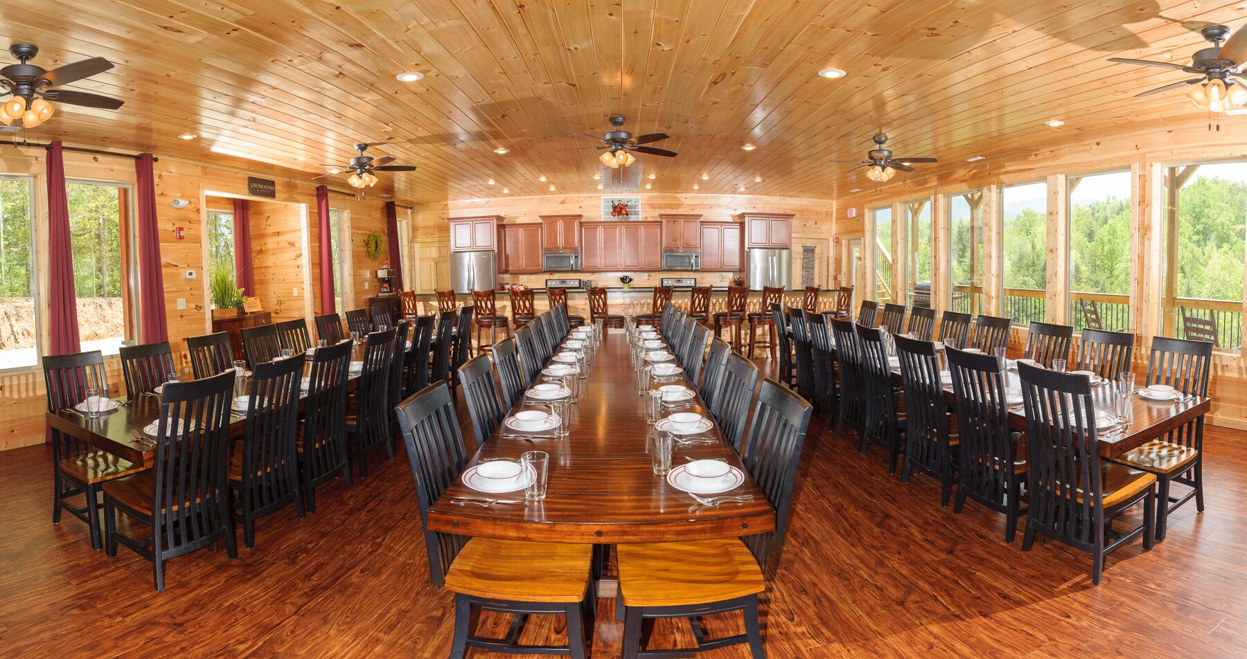 Une vaste salle à manger en bois, avec de nombreuses tables dressées pour un repas, équipée de ventilateurs de plafond et de grandes fenêtres donnant sur l’extérieur.